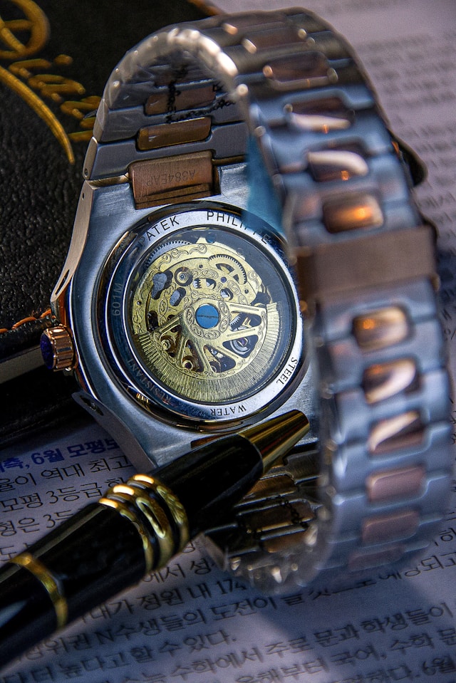 Close-up of a Patek Philippe showing its intricate movement through a transparent caseback, surrounded by a silver metal bracelet and a black pen resting on paper with text.