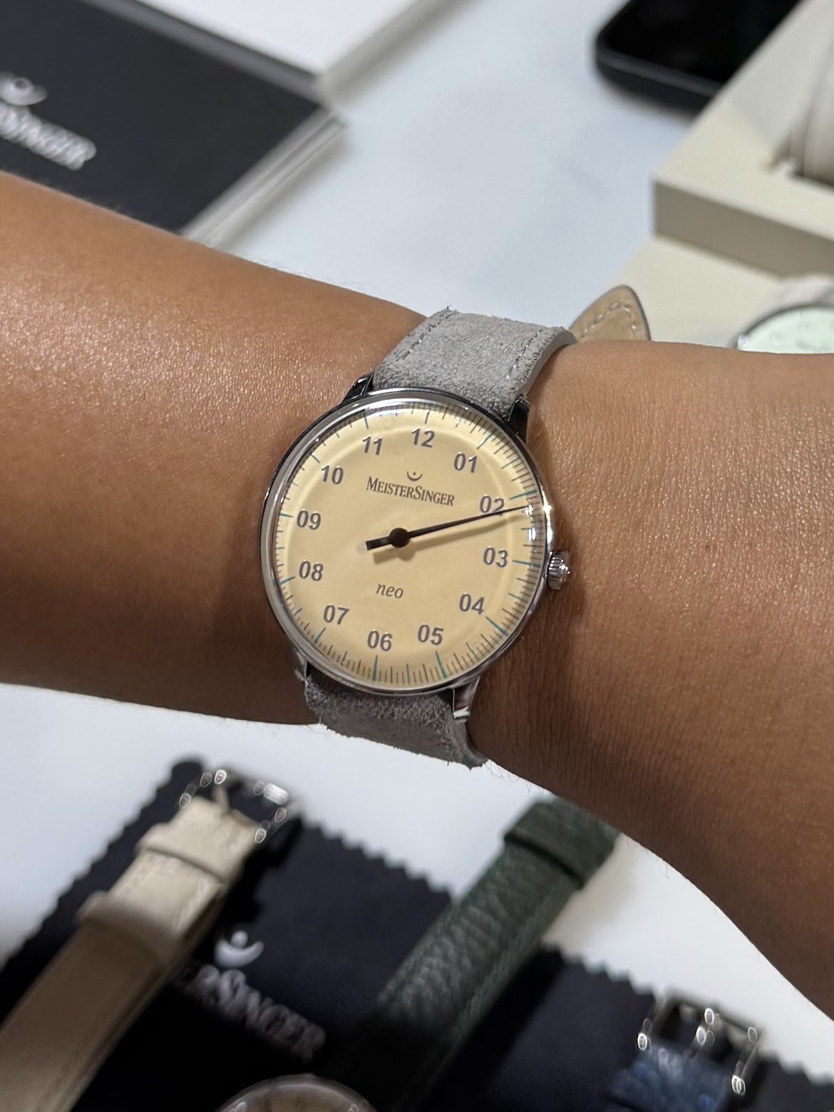Close-up of a hand wearing a minimalist watch with a light-colored dial and silver accents, surrounded by other watches on a display table at a watch fair.