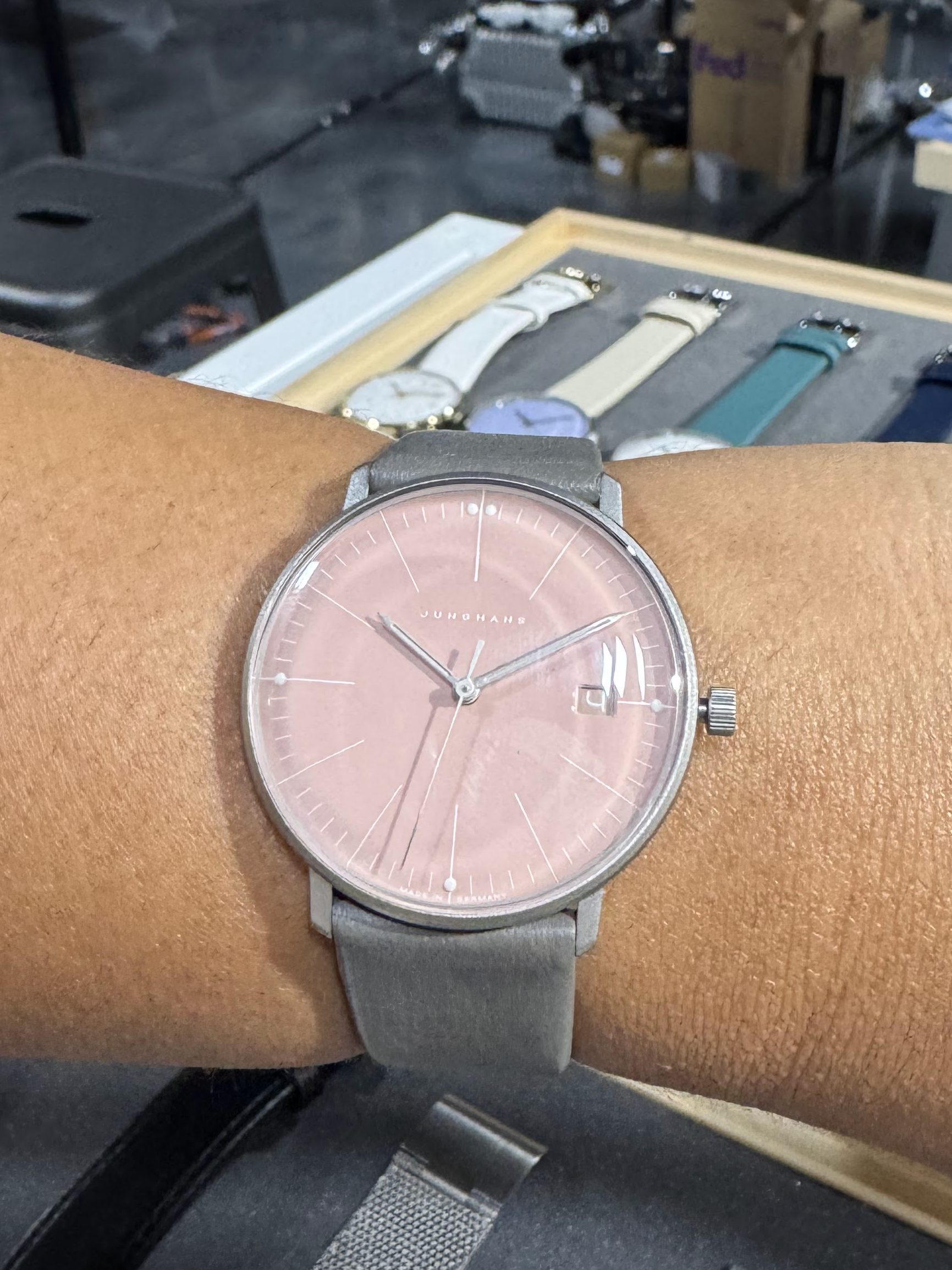 A close-up view of a person's wrist wearing a Junghans Max Bill watch with a pastel pink dial and gray strap, set against a backdrop of various watch straps displayed on a table.