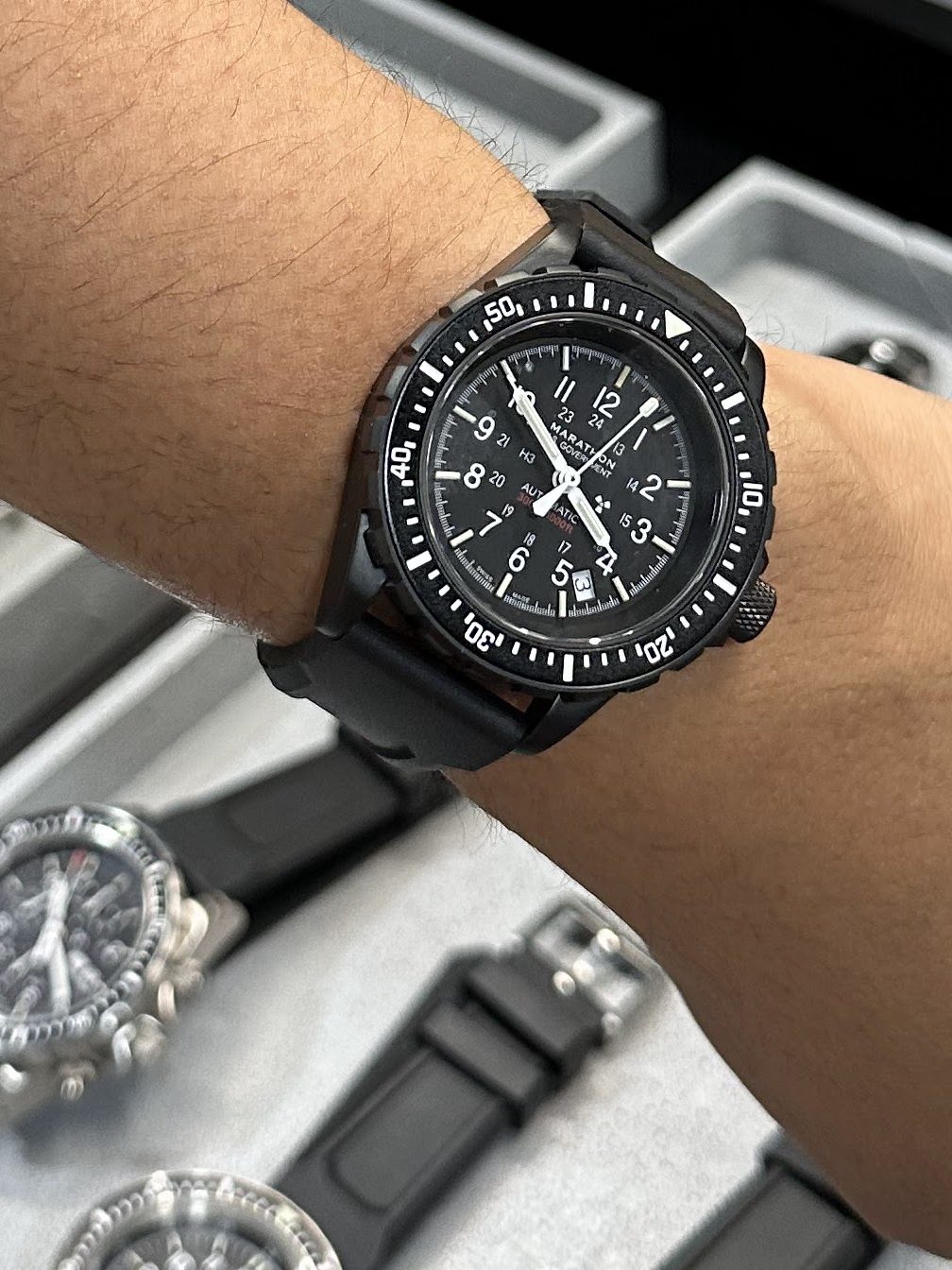 A close-up of a wrist wearing a black chronograph watch, displayed alongside various other watches on a table at the Windup Watch Fair.